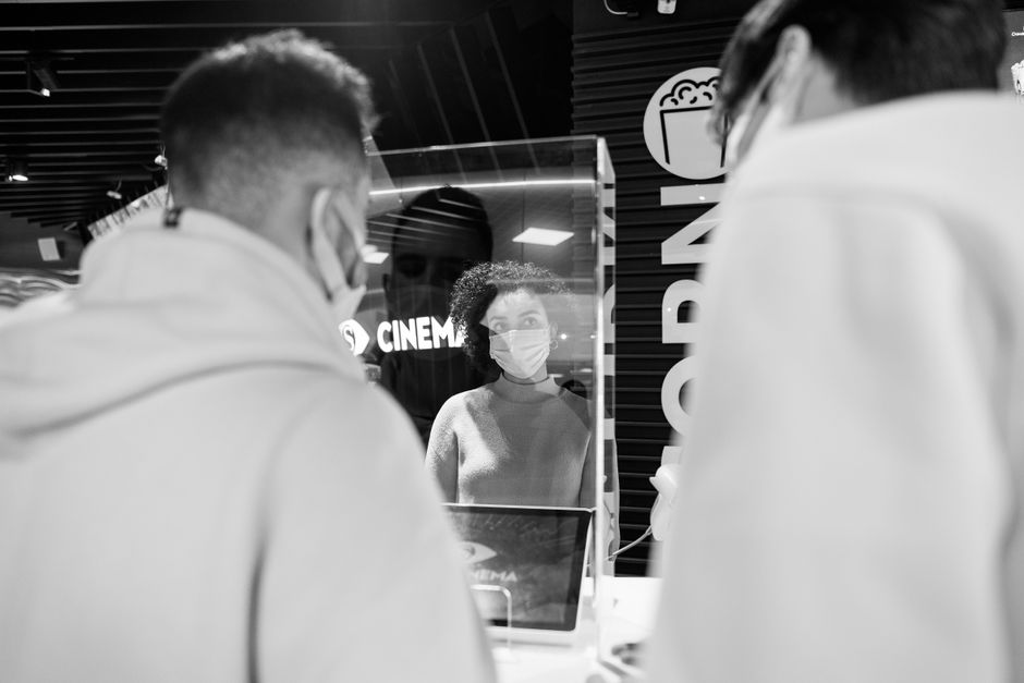 Black and white image of cinema ticket counter with masked individuals during pandemic.